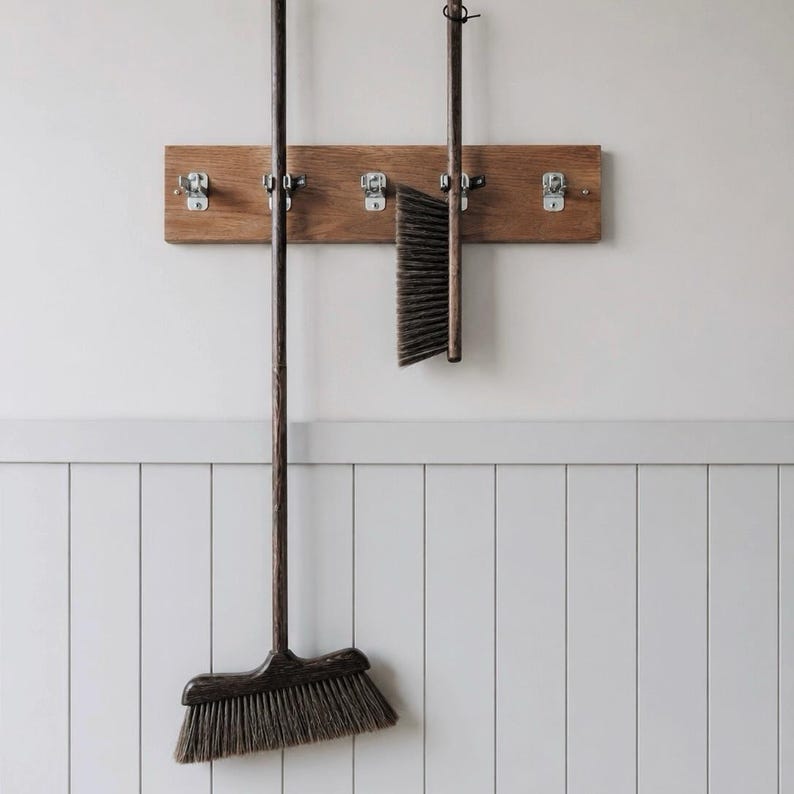 May include: A rectangular wooden wall rack with five silver metal hooks holding two dark brown wooden cleaning brushes. The long-handled broom and the shorter hand brush feature dark bristles and are suspended vertically from the wall-mounted rack.