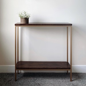 May include: A modern console table with a dark wood top and lower shelf. The frame is a gold color. A small, round, brown pot with white flowers sits on the top shelf. The table is approximately 30 inches long, 12 inches wide, and 30 inches tall.