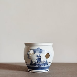 May include: White ceramic planter with a blue and white windmill design. The planter has two large circular holes and a dark blue rim. The design features a windmill, boat, and clouds. A decorative piece for plants or home decor.