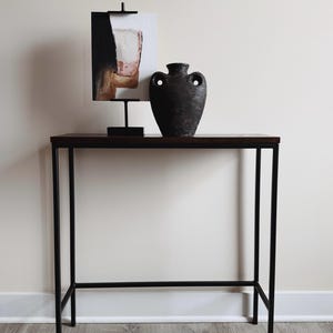 May include: A tall, dark brown, textured ceramic vase with two side handles sits on a rectangular console table with a dark wood top and black metal frame. Next to the vase is an abstract painting in a black metal stand with a black base.