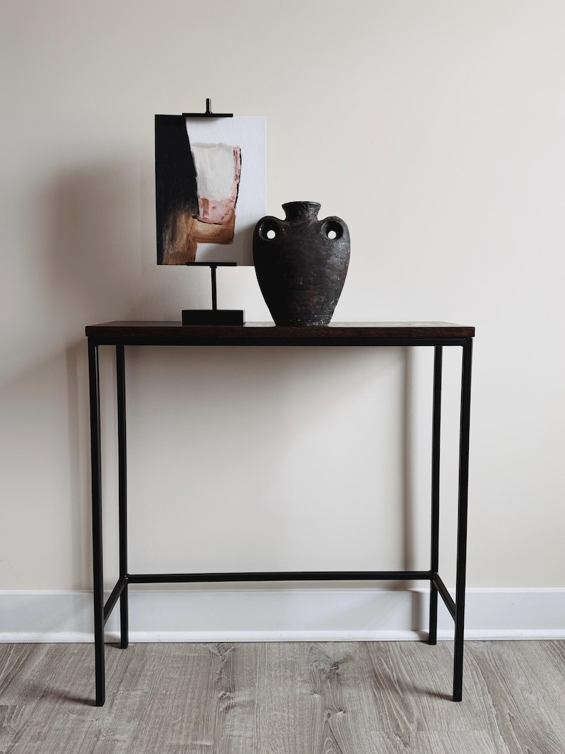 May include: A tall, dark brown, textured ceramic vase with two side handles sits on a rectangular console table with a dark wood top and black metal frame. Next to the vase is an abstract painting in a black metal stand with a black base.