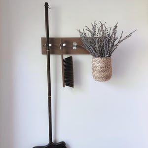 May include: A rectangular wooden wall-mounted rack with three silver metal hooks. A long-handled dark wood broom, a handheld brush with dark bristles, and a woven seagrass basket filled with dried lavender sprigs hang from the hooks.