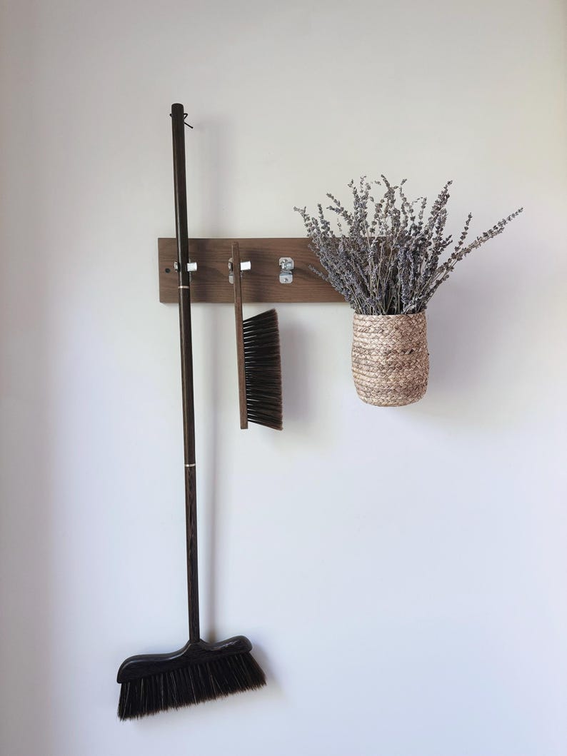 May include: A rectangular wooden wall-mounted rack with three silver metal hooks. A long-handled dark wood broom, a handheld brush with dark bristles, and a woven seagrass basket filled with dried lavender sprigs hang from the hooks.