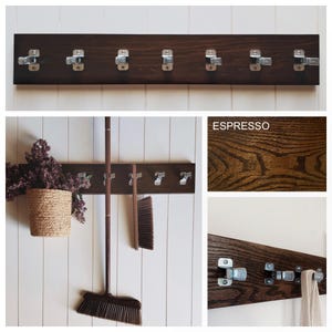 May include: A dark brown espresso-stained wooden wall rack featuring seven silver metal spring-loaded clips for hanging items. The rectangular board displays a visible wood grain texture and is shown holding a broom and a cloth.