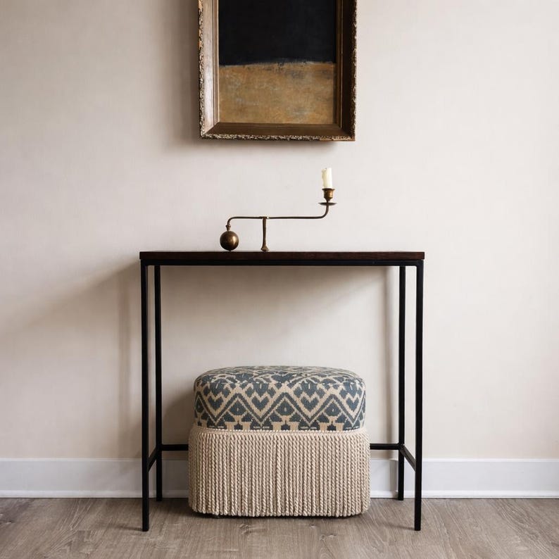 May include: A rectangular console table with a dark wood top and black metal frame. On top sits a brass candle holder. Below the table is an upholstered ottoman featuring a blue and white geometric pattern with a thick, cream-colored rope fringe.
