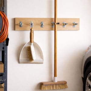 May include: A light-coloured wooden wall-mounted rack featuring six silver metal spring-loaded clips. A white plastic dustpan with a wooden handle and a broom with a long wooden handle are secured to the rack by the metal clips.