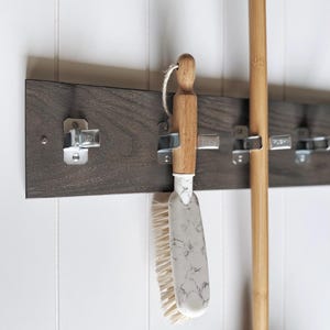 May include: A dark brown wooden wall-mounted organiser rack featuring four silver metal spring-loaded clips with the word PUSH embossed on each. A cleaning brush with a light wood handle and a white marble-patterned head hangs from one clip.