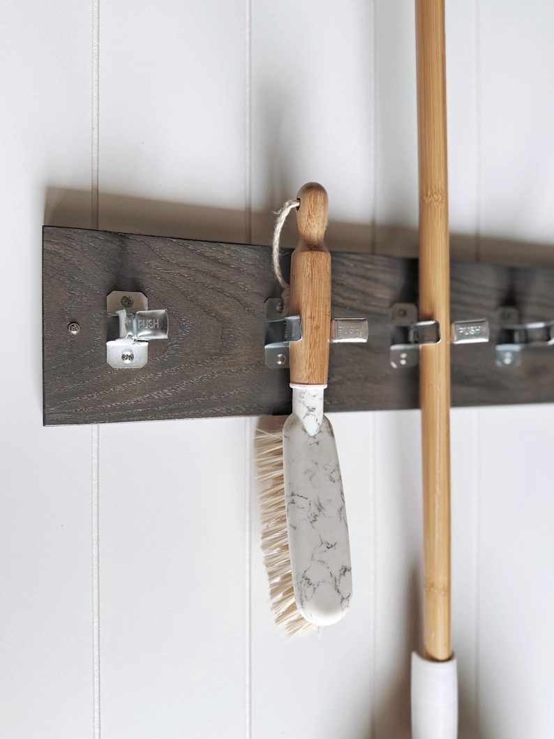 May include: A dark brown wooden wall-mounted organiser rack featuring four silver metal spring-loaded clips with the word PUSH embossed on each. A cleaning brush with a light wood handle and a white marble-patterned head hangs from one clip.