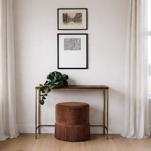 May include: A brown velvet cylindrical ottoman with a thick fringe base sits beneath a slim console table with a dark wood top and gold metal frame. Two framed black and white prints hang on the wall above, accompanied by a green trailing plant.