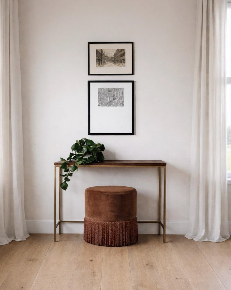 May include: A brown velvet cylindrical ottoman with a thick fringe base sits beneath a slim console table with a dark wood top and gold metal frame. Two framed black and white prints hang on the wall above, accompanied by a green trailing plant.