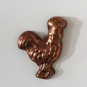 May include: A copper-colored rooster-shaped mold for baking. The mold is detailed with a textured surface and has a slightly raised tail.