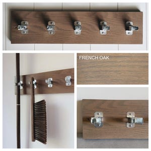 May include: A rectangular brown French oak wall-mounted organiser featuring five silver metal spring-loaded clips. The wood grain is visible, and the clips are secured with screws. One clip holds a dark-bristled brush, and another holds a long handle.