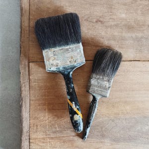 May include: Two paint brushes with black bristles and worn handles rest on a wooden surface. The larger brush has a black handle with yellow stripes and the text "Gold Stripe". The smaller brush has the text "WORKMASTER".