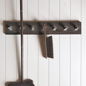 May include: A dark brown wooden wall-mounted rack featuring seven silver metal spring-loaded clips. A long-handled broom and a hand brush, both with dark brown wooden handles and dark bristles, are secured to the rack by the metal clips.