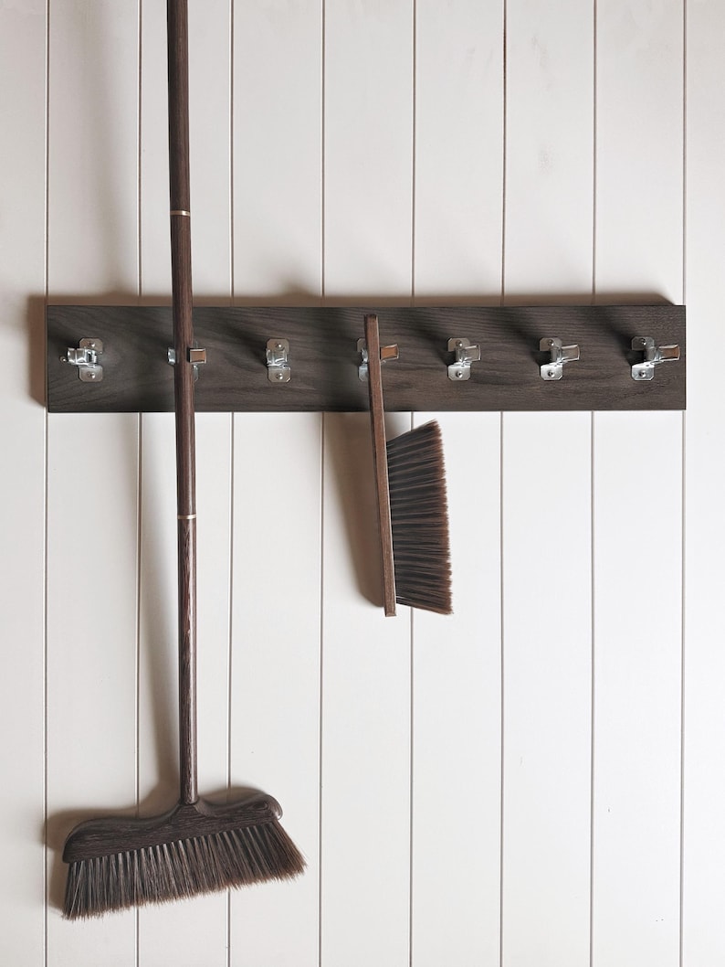 May include: A dark brown wooden wall-mounted rack featuring seven silver metal spring-loaded clips. A long-handled broom and a hand brush, both with dark brown wooden handles and dark bristles, are secured to the rack by the metal clips.