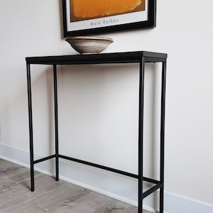 May include: A tall, narrow console table featuring a slim black metal frame with four straight legs and a rectangular black top. A shallow, round ceramic bowl with a neutral, multi-toned finish sits centered on the surface of the table.