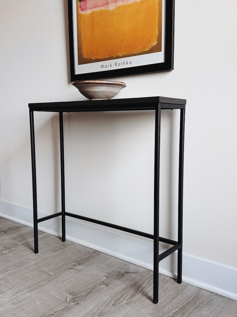 May include: A tall, narrow console table featuring a slim black metal frame with four straight legs and a rectangular black top. A shallow, round ceramic bowl with a neutral, multi-toned finish sits centered on the surface of the table.