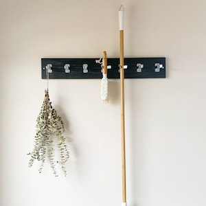 May include: A dark wood wall-mounted rack with six silver metal clips. A dried eucalyptus bundle hangs from the first clip, a white and wood scrub brush hangs from the fourth, and a long-handled broom with a wood handle hangs from the fifth.