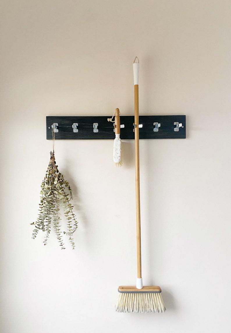May include: A dark wood wall-mounted rack with six silver metal clips. A dried eucalyptus bundle hangs from the first clip, a white and wood scrub brush hangs from the fourth, and a long-handled broom with a wood handle hangs from the fifth.