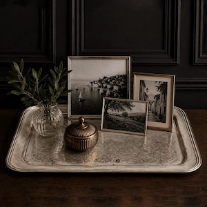 May include: A large rectangular silver-toned metal tray with an ornate engraved border. On the tray sits a clear ribbed glass vase with green leaves, a round lidded bronze-toned container with vertical ridges, and three framed black and white photos.