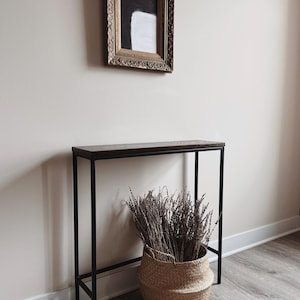 May include: A rectangular console table with a dark wood top and a thin black metal frame. A woven seagrass basket filled with dried lavender stems sits on the floor beneath the table. Above the table hangs a framed painting with a gold border.