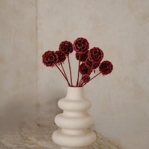 Può includere: Un vaso in ceramica bianco sporco opaco con una silhouette impilata, bulbosa e a gradini contenente un bouquet di fiori secchi color bordeaux intenso a forma di globo su steli sottili e abbinati che si estendono dal collo stretto.