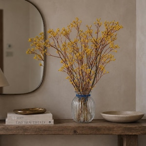 Könnte beinhalten: Eine klare, vertikal gerippte Glasvase mit einem Strauß aus dünnen, braunen Zweigen mit kleinen gelben Blüten. Die Vase ist mit einer dünnen blauen Kordel um den Hals gebunden und steht auf einer Holzoberfläche neben Büchern und Schalen.