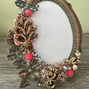 May include: Oval-shaped, ornate picture frame with a bronze finish. The frame is embellished with a variety of decorative elements, including a dragonfly, flowers, pearls, and rhinestones. The frame is designed to hold a photo.