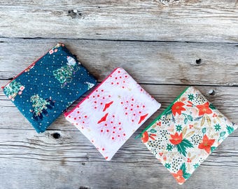 May include: Three quilted zipper pouches with holiday-themed patterns. One is navy blue with Christmas trees, another is white with red trees, and the third is cream with red flowers and green accents. Each pouch has a different colored zipper.