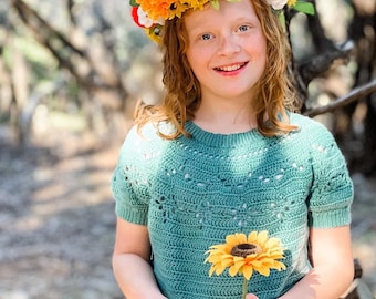 May include: A young girl with red hair wearing a crocheted top and a flower crown made of orange, yellow, and red flowers. She is holding a single sunflower.