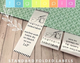 Könnte beinhalten: Zwei gefaltete Etiketten aus weißem Satin mit schwarzem Text und einer Grafik mit einer Nähnadel und einem Faden. Der Text auf den Etiketten lautet "tagtopia folded labels" und "machine wash tumble dry no bleach no iron 1x3".