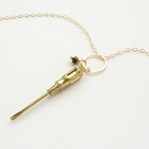 Könnte beinhalten: Eine goldene Halskette mit einem Schraubendreher-Anhänger und einer kleinen schwarzen Perle. Die Kette hat eine zarte Kette und einen runden Anhänger.