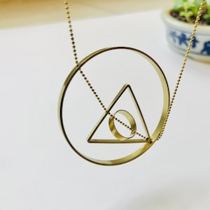 May include: A gold necklace with a geometric pendant featuring a circle and a triangle with a smaller circle inside. The pendant is suspended from a delicate chain.
