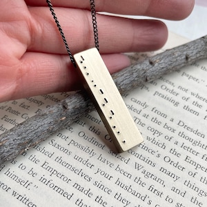 May include: A rectangular gold-toned metal pendant suspended from a thin black chain. The pendant features a vertical column of small, raised dots and dashes arranged in a Morse code pattern on its front surface, creating a textured appearance.