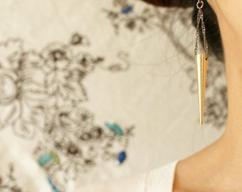 Può includere: Un orecchino a forma di cono dorato con una catena d'argento appesa a un gancio d'argento. L'orecchino è indossato nell'orecchio sinistro di una persona con i capelli castani.