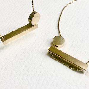 May include: Two gold necklaces with geometric pendant designs. One necklace features a round pendant and a long, thin bar pendant. The other necklace features a round pendant and a shorter, thicker bar pendant.