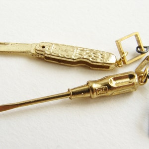 Könnte beinhalten: Ein Paar goldfarbene Ohrringe mit einem Miniatur-Taschenmesser und einem Miniatur-Schraubendreher. Jeder Ohrring hat einen quadratischen Anhänger und einen schwarzen Drahtbügel.