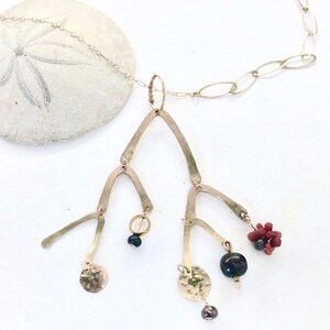 Puede incluir: Un collar de oro con un largo colgante. El colgante está hecho de múltiples piezas de metal curvadas que se asemejan a ramas. El colgante está adornado con cuentas negras y rojas, y un pequeño anillo de oro.