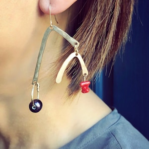 May include: Artisan earrings with a unique, asymmetrical design. The earrings feature a silver-toned, curved bar, a gold-toned arch, a dark gray pearl, and red coral accents. The earrings are suspended from a simple ear wire.