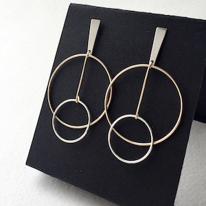 Puede incluir: Un par de pendientes de aro de oro y plata con un círculo más pequeño dentro del círculo más grande. Los pendientes están en un expositor negro.