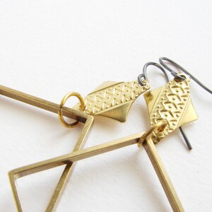 May include: Gold geometric earrings with a square frame and a patterned diamond-shaped pendant. The earrings have a black wire hook.