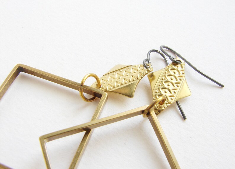 May include: Gold geometric earrings with a square frame and a patterned diamond-shaped pendant. The earrings have a black wire hook.