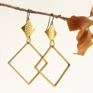 May include: A pair of gold-toned earrings with geometric square shapes. The earrings are suspended from a branch with brown leaves.