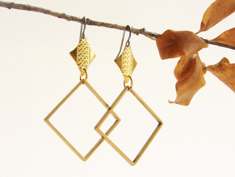 May include: A pair of gold-toned earrings with geometric square shapes. The earrings are suspended from a branch with brown leaves.