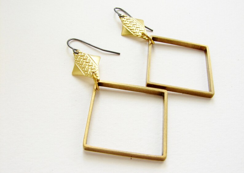May include: A pair of gold-toned earrings with a geometric square design. Each earring features a small, textured diamond-shaped piece at the top.