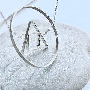 May include: A silver necklace with a geometric pendant featuring a circle, triangle, and square. The pendant is suspended from a delicate silver chain.