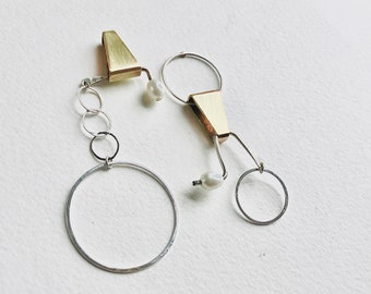 Könnte beinhalten: Ein Paar Ohrringe aus Gold und Silber mit geometrischem Design und Perlenakzenten. Die Ohrringe haben eine dreieckige Form mit einem daran befestigten Kreis. Die Ohrringe hängen an einem kleinen Ring.
