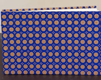 May include: A rectangular notebook with a vibrant blue cover featuring a repeating pattern of gold circular motifs and small four-petaled flower shapes. The spine side shows visible thread stitching binding the pages together in a horizontal orientation.