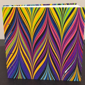 May include: A square-bound book featuring a vibrant marbled cover with a repeating chevron pattern in bright shades of yellow, hot pink, lime green, purple, and blue. The spine edge shows visible blue thread stitching along the binding.
