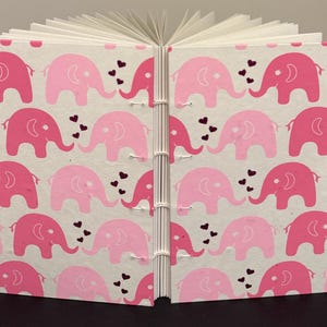 May include: A handmade journal with a pink elephant pattern. The cover features a repeating design of pink elephants and small heart shapes on a white background. The journal is bound with white thread.